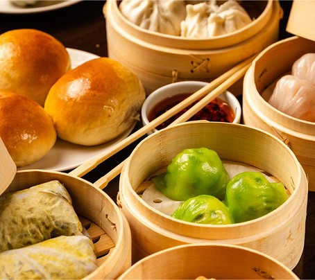 An assortment of dim sum in bamboo steamers, including green dumplings, buns, and a side of chili sauce.