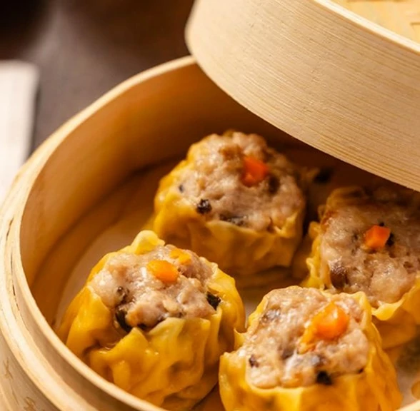 A bamboo steamer holds four golden siu mai dumplings, each topped with a small orange carrot piece, showcasing their savory filling.
