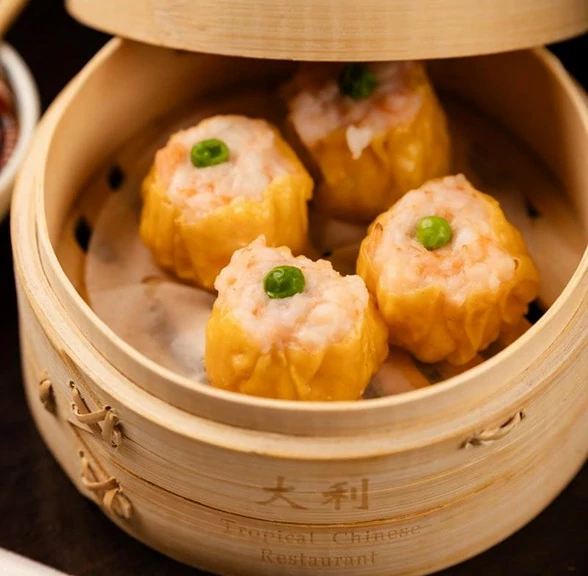 Four golden siu mai dumplings with shrimp filling, garnished with green peas, served in a bamboo steamer.