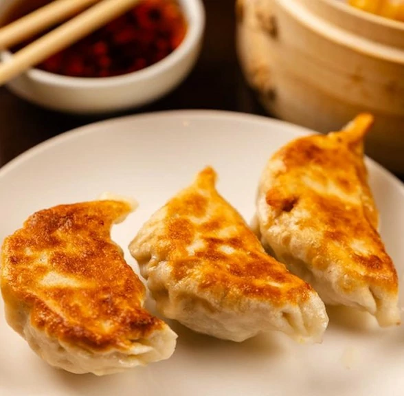 Three golden-brown dumplings on a white plate, accompanied by dipping sauce and chopsticks, with a bamboo steamer in the background.