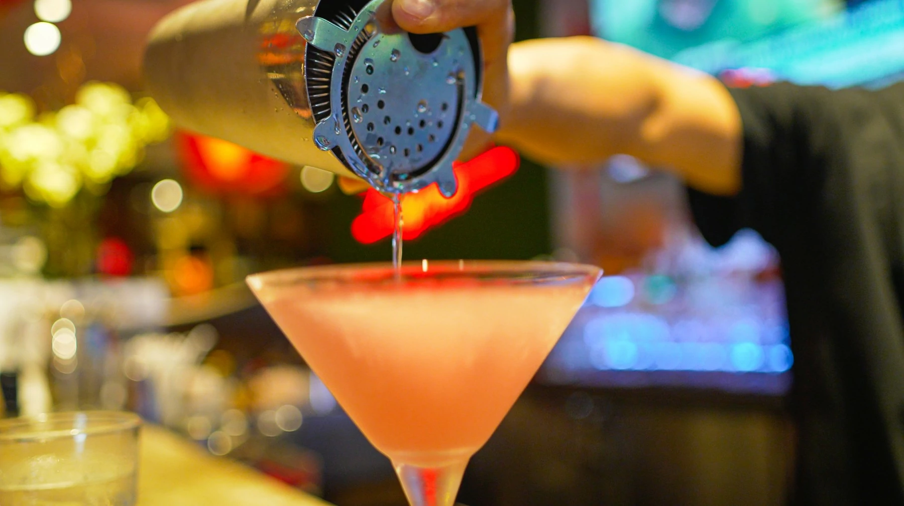 A bartender pours a vibrant pink cocktail from a shaker into a martini glass at a lively bar, with colorful blurred lights in the background highlighting the drink-making process.